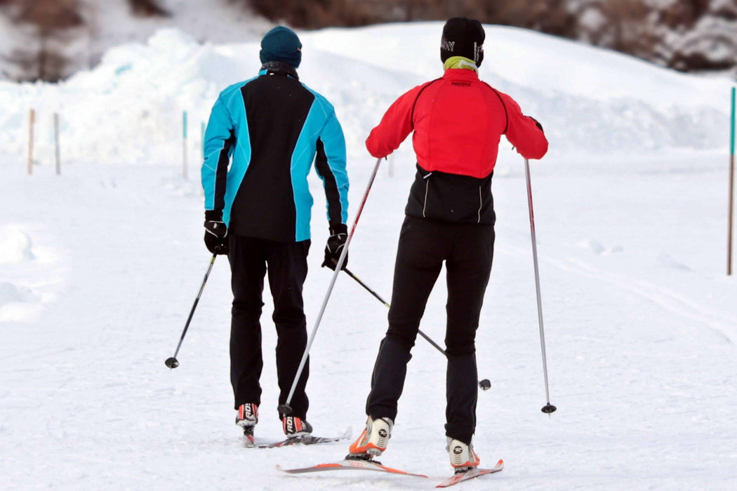 Two cross country skiers moving away from the camera. Skier on the right  is wearing  a red jacket and black ski pants and the other is in a blue and black jacket with black snow pants.  Wearing the Phonak Stick 'n' Stay Hearing Aid Stickers (30 pair) 098-0353 Phonak