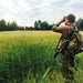 Serenity Choice Reusable Hearing Protection for HUNTING & SHOOTING Earplugs.  Hunter standing in a green field of wheat looking through binoculars across the field to a line of evergreen trees.  