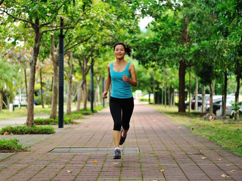 Woman running on a paved path in a park with trees and cars in the background