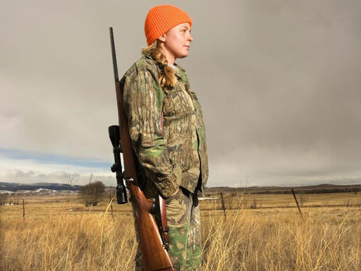 Person in camouflage clothing and orange beanie holding a rifle in an open field.