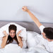 A couple in bed are stressed from sounds on the other side of the wall.  The woman covers her ears with a pillow and the man pounds on the wall with his fist.  They would benefit from using the Serenity Choice Reusable Hearing Protection for SLEEP Earplugs.