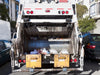 Back of a garbage truck on a city street with buildings and cars in the background.