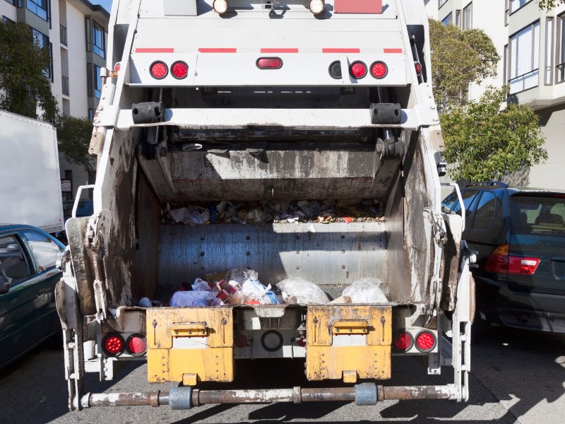 Back of a garbage truck on a city street with buildings and cars in the background.