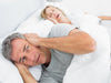 A man covering his ears with a woman sleeping in bed behind him, snoring. 