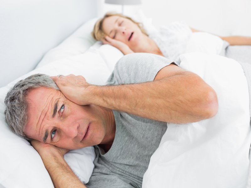 A man covering his ears with a woman sleeping in bed behind him, snoring. 
