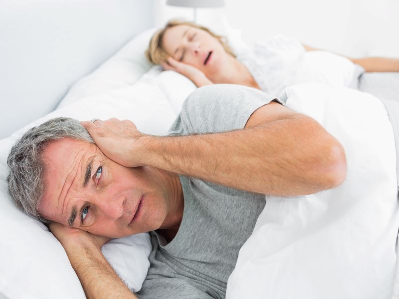 A man covers his hears with his hands as his wife snores behind him in bed.  He would be sleep if he had not forgotten his Serenity Choice Reusable Hearing Protection for SLEEP Earplugs.