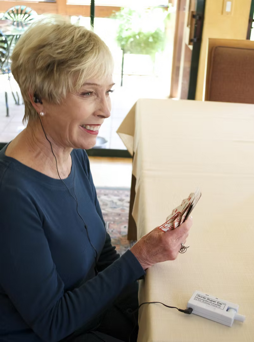 A woman is holding a hand of playing cards and using the SuperEar® Model SE5000 Compact Personal Sound Amplifier with earbuds. 