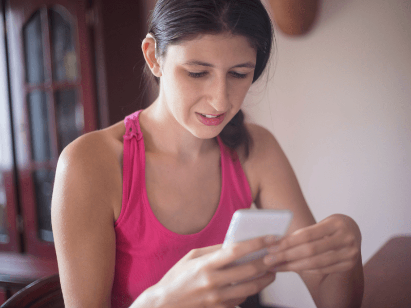 A young woman wearing hearing aids and  in a pink tank top sits on her cell phone.  She uses the Phonak Stick 'n' Stay Hearing Aid Stickers (30 pair) 098-035 Adhesives. 