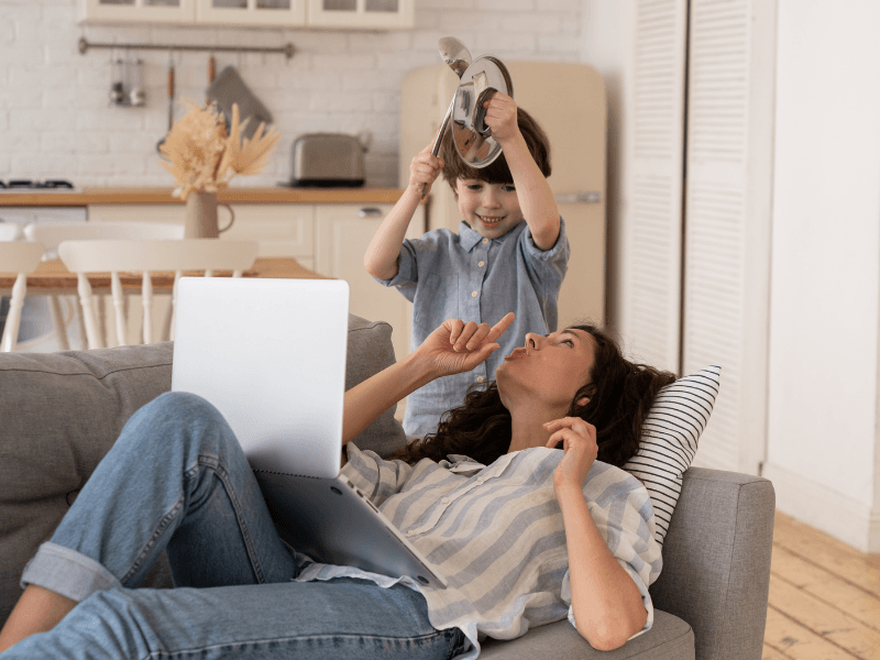Serenity Choice Reusable Hearing Protection can be used in many situations.  In this photo a mother is laying on the couch with her laptop holding her pointer finger to her lips as she looks at her young son.  The son is holding a pot lid and ladle above his head. 