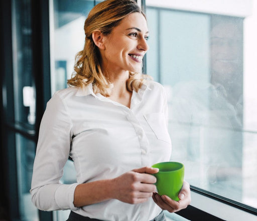 Serenity Choice Reusable Hearing Protection can be used in an office environment.  A blonde woman dressed in a white button up blouse stands leaning against a window looking out.  She holds a green mug  in her hands as she smiles and looks out the window. 