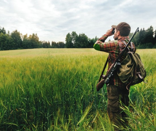 Serenity Choice Reusable Hearing Protection for HUNTING & SHOOTING Earplugs.  Hunter standing in a green field of wheat looking through binoculars across the field to a line of evergreen trees.  