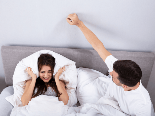 A couple in bed are stressed from sounds on the other side of the wall.  The woman covers her ears with a pillow and the man pounds on the wall with his fist.  They would benefit from using the Serenity Choice Reusable Hearing Protection for SLEEP Earplugs.