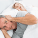 A man covers his hears with his hands as his wife snores behind him in bed.  He would be sleep if he had not forgotten his Serenity Choice Reusable Hearing Protection for SLEEP Earplugs.