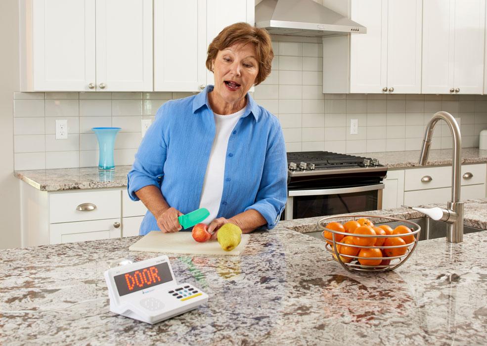 Sonic Alert HomeAware™ Wireless Doorbell HA360DB2-1 sends a wireless message to the Main Receiver to display the red word DOOR on a black screen.  The Main receiver sits on a kitchen counter top as a woman cuts up an apple on a cutting board.  